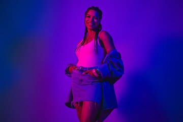 Lovely smiling woman in stylish denim outfit posing under vibrant neon lights in a modern studio