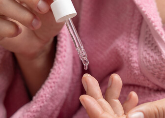 Young woman in pink bathrobe dropping essential oil on a finger and holding a pipette closeup