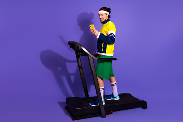 Young athletic man on a treadmill using a cellphone in vibrant sporty attire against a vivid purple...