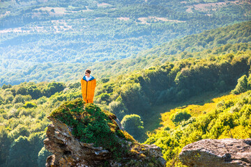 Man in sleeping bag standing on cliff, looking at sunrise over mountain valley