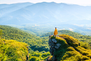Man wrapped in a blanket standing on mountain cliff, admiring panoramic view of forest valley and distant hills at sunset