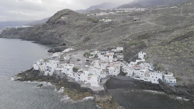 Drone orbits to the left in wide shot of coastal town with white buildings on cloudy day in El Varadero, Tenerife, Canary Islands, Spain