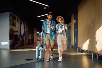 Happy couple using mobile phone while walking through departure terminal at station.