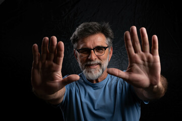 Mature handsome man smiling with outstretched hands toward camera, black background, chiaroscuro...