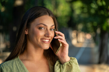 Smiling young woman talking on smartphone outdoors, close up portrait looking at camera in park or...