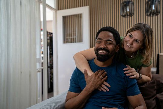 Diverse happy couple smiling and looking together into the future in cozy living room, love and optimism for bright days