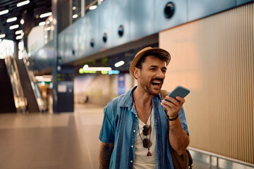 Happy man recording voice message on smart phone while walking through airport terminal.
