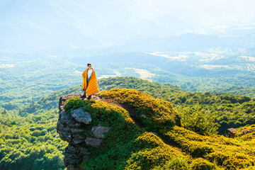 Man wrapped in a blanket standing on mountain cliff, admiring panoramic view of forest valley and distant hills at sunset