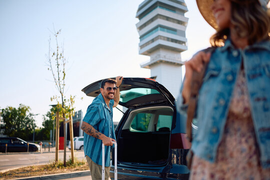 Happy man traveling with his girlfriend and taking suitcase out of the car trunk at airport parking lot.