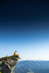 Man in yellow sleeping bag sitting on cliff edge, looking out over mountain landscape under clear sky