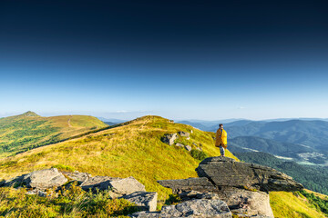 Man wrapped in a blanket standing on mountain cliff, admiring panoramic view of forest valley and distant hills at sunset