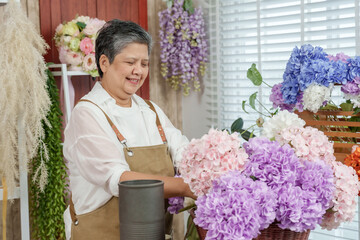 Asian elderly female florist arranging artificial flowers with care and passion at flower shop...