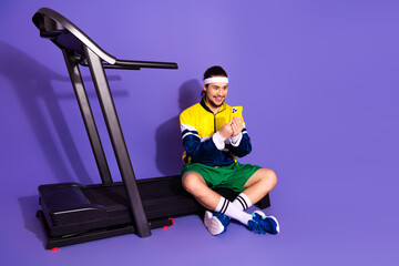 Young athlete in retro-inspired activewear seated near a treadmill using a smartphone against a...