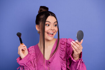 Happy young woman holding a makeup brush and mirror while wearing a stylish pink ruffled blouse...