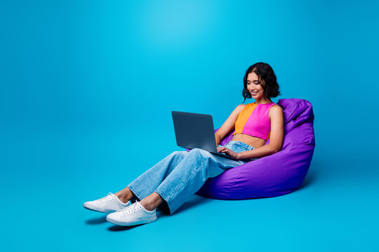 Cheerful woman using a laptop while sitting on a purple bean bag in a vibrant blue setting for lifestyle and leisure