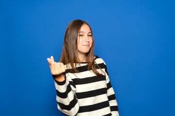A beautiful Hispanic teenager in her 15s wearing a striped sweater posing isolated against a blue background. Lifestyle concept. Rubbing her fingers together, gesturing for money
