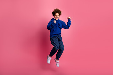 Energetic boy jumping joyfully in a blue hoodie against a bright pink background showing vibrant...