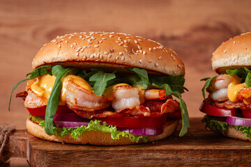 two shrimp burgers and buns with sesame seeds, bacon, lettuce, tomatoes and onions,on a wooden board, rustic style,