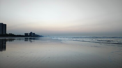 Nascer do sol dourado na Praia Grande, SP. Silhuetas urbanas refletidas na orla calma sob luz...