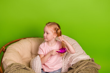 Young girl in casual clothes brushing hair, seated on a cozy chair with a green background, reflecting care