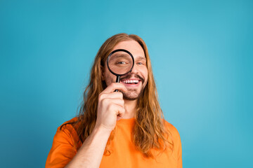 Young man with long hair holding a magnifying glass playfully smiling in a joyful blue background...