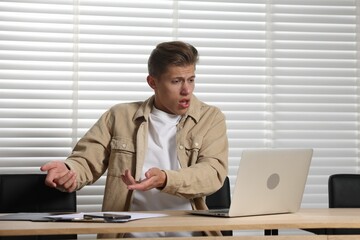 Young man with papers complaining to someone via laptop at wooden table indoors