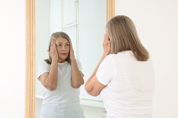 Beautiful senior woman looking at herself in mirror indoors