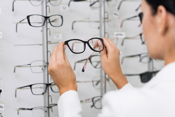 Attractive Caucasian young brunette woman in white suit choosing eyeglasses on showcase for purchase at an optical store. Lifestyle, optometry, eyesight, assortment of glasses