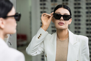 Elegant Caucasian young brunette woman in white suit trying on sunglasses looking in mirror for purchase at optical store. Lifestyle, optometry, eyesight, eye protection