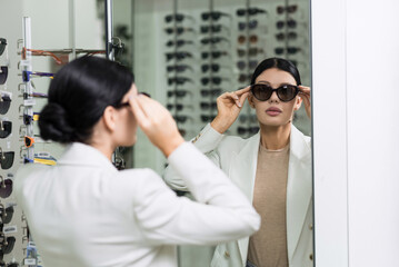 Elegant Caucasian young brunette woman in white suit trying on sunglasses looking in mirror for purchase at optical store. Lifestyle, optometry, eyesight, eye protection