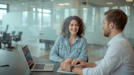 Job interview in office boardroom, woman with curly hair, wearing a denim shirt, is seated at a long dark conference table in an office, laughing while looking at a man across from her