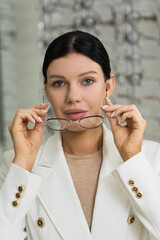 Attractive Caucasian young brunette woman in white suit trying on and choosing eyeglasses for buying at an optical store. Lifestyle, optometry, eyesight, assortment of glasses