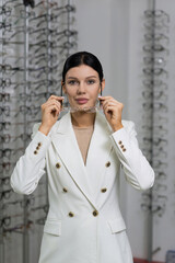 Attractive Caucasian young brunette woman in white suit trying on and choosing eyeglasses for buying at an optical store. Lifestyle, optometry, eyesight, assortment of glasses
