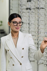 Attractive Caucasian young brunette woman in white suit trying on and choosing eyeglasses for buying at an optical store. Lifestyle, optometry, eyesight, assortment of glasses