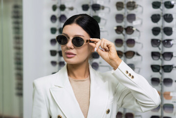 Attractive Caucasian young brunette woman in white suit chooses eyeglasses to buy at an optical store. Lifestyle, optometry, eyesight, assortment of glasses