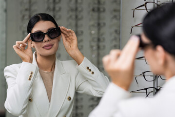 Elegant Caucasian young brunette woman in white suit trying on sunglasses looking in mirror for purchase at optical store. Lifestyle, optometry, eyesight, eye protection.