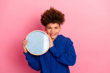 Smiling boy in blue hoodie holding large clock against pink wall background, perfect representation...