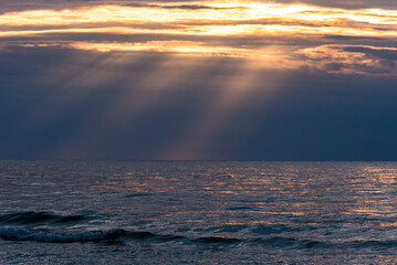 Obraz premium Calm sea under a cloudy sky at sunset. Sunlight filters through the clouds, creating rays over the water. 