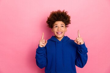 Joyful teenager wearing a stylish blue hoodie pointing upwards against a vibrant pink background,...