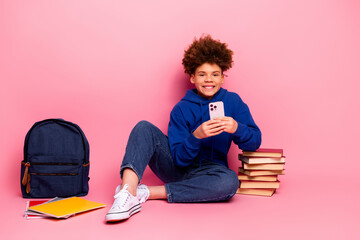Smiling teenage boy holding smartphone with stack of book near pink background in stylish hoodie
