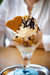 Close-up of a creamy ice cream sundae topped with whipped cream, chocolate syrup, crunchy cookie bites, and a heart-shaped waffle in a glass cup. Elegant and indulgent dessert.