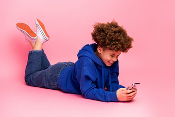 Young boy in blue hoodie using smartphone on pink background showing cheerful expression and modern...