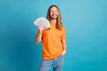 Cheerful man holding cash fan with a stylish look in a vibrant orange shirt over a bright blue...