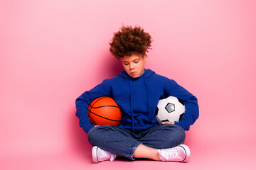 Young boy in a blue hoodie sitting with basketball and soccer ball on a pink background showcasing sports and choice