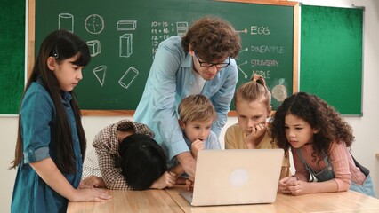 Caucasian teacher and diverse student looking at laptop at classroom. Cute multicultural children learning about coding program or generated program by using software while teacher teaching. Pedagogy.