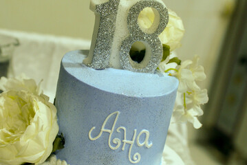A two-tiered blue and white birthday cake decorated with a pearl ribbon and white and yellow flowers with the female name Ana written in Cyrillic script