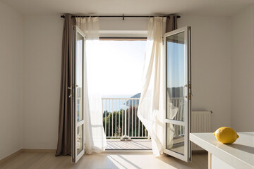 Open balcony door with a white curtain blowing in a gentle breeze, creating a bright, airy, and peaceful atmosphere in a minimalist room with a single fresh lemon on a table