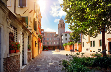 Venice, Italy. Venetian old town courtyard, Antique well between colorful buildings and a bell tower. Sunny Venezia street with green trees and medieval Italian architecture.