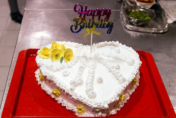 White heart shaped birthday cake. Decorated with yellow flowers and white whipped cream with the inscription Happy Birthday