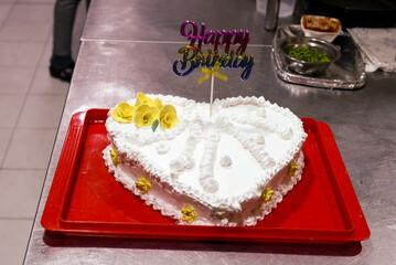 White heart shaped birthday cake. Decorated with yellow flowers and white whipped cream with the inscription Happy Birthday